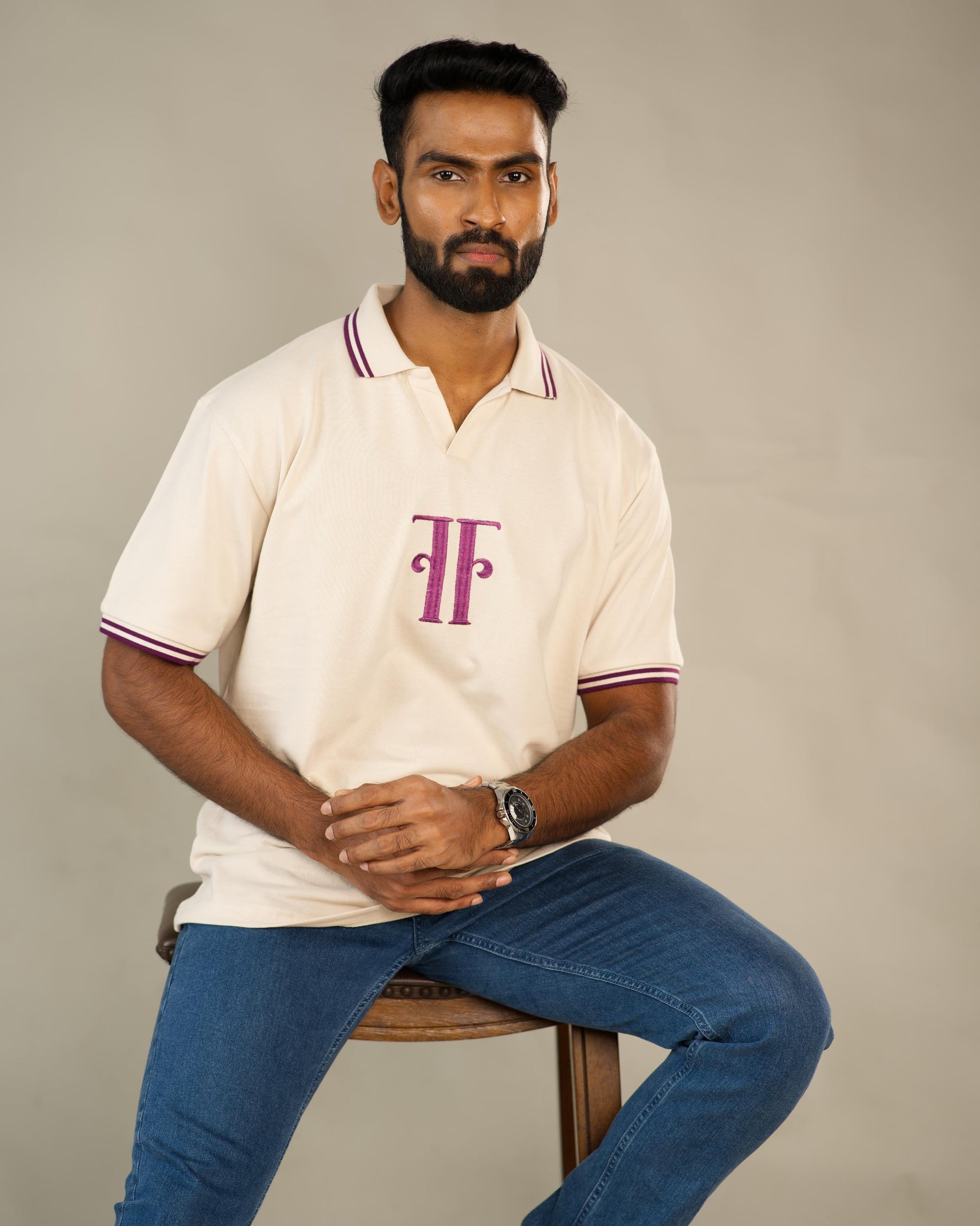 Man wearing a white polo shirt with a logo and blue jeans sitting on a chair against a plain background - Image 1