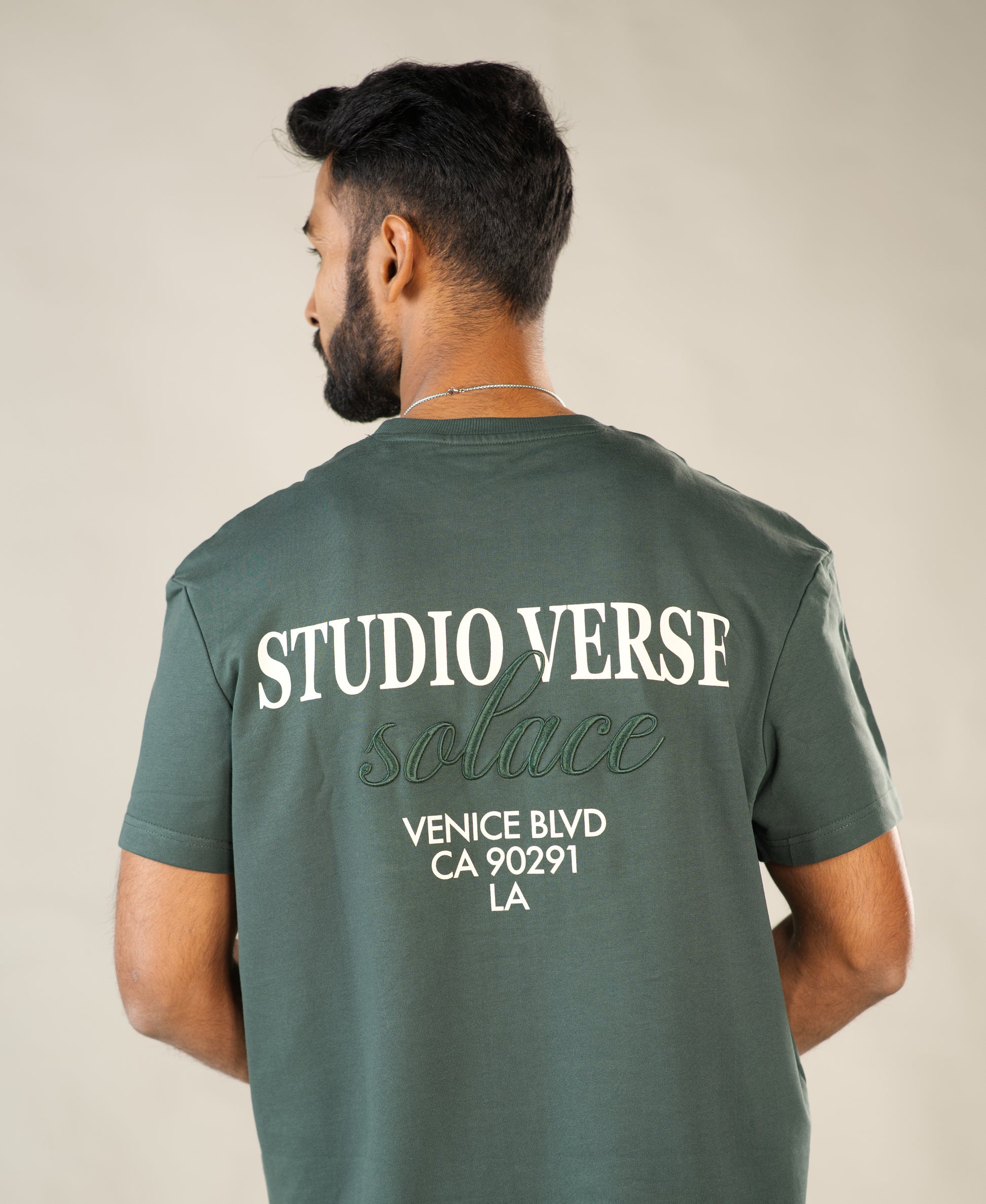Man wearing a green t-shirt with 'Studio Verse Solace' text on the back against a beige background - Image 2